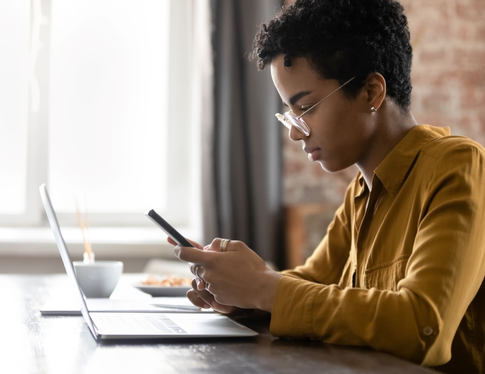 Woman sits at desk looking at smartphone. Open laptop computer is in front of her.