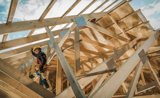 A man in a hard hat works on building a roof, wooden beams are all around him. 
