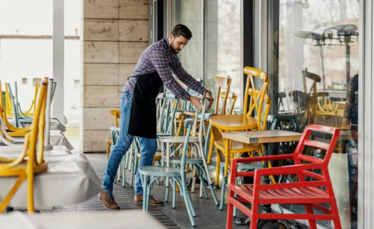 Waiter in a plaid shirt and apron stacks multicolored patio chairs against an outside glass wall.
