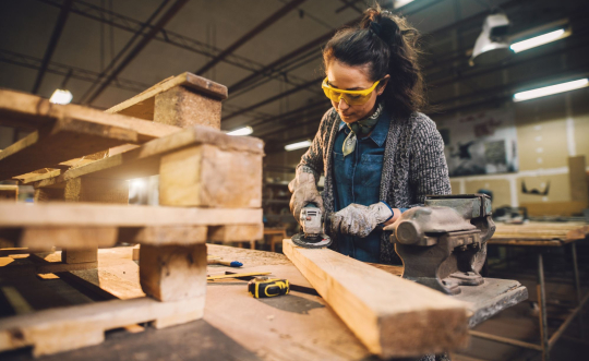 A middle-aged woman wearing a denim shirt, sweater, safety glasses and gloves cuts a 2x4 with a powersaw.  Cut wood is stacked carefully beside her.