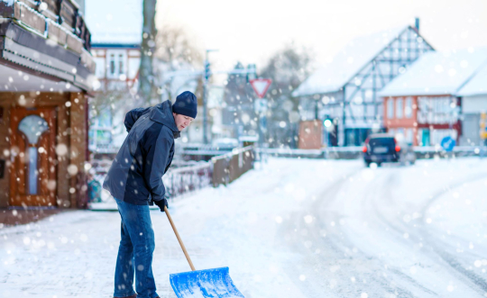 Man in gray and black coat with ski cap shovels the cobblestone walkway in front of a business.