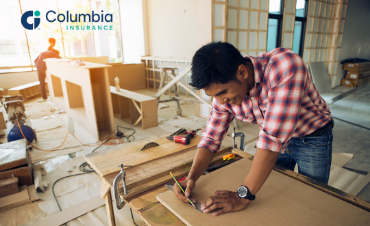 A contractor in a red plaid shirt marks a measurement on a sheet of particle board.