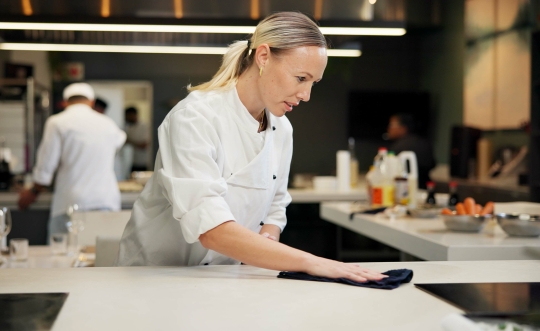 A blond female in a white chef coat wipes up the counter of a commercial kitchen. 
