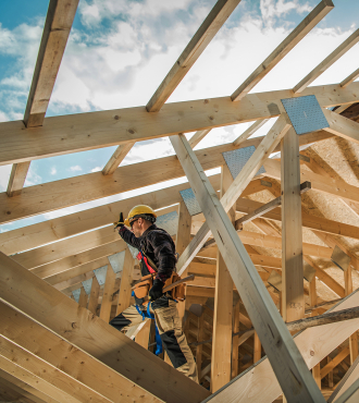 A man in a hard hat works on building a roof, wooden beams are all around him. 