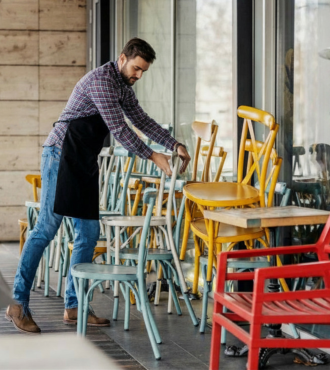 Waiter in a plaid shirt and apron stacks multicolored patio chairs against an outside glass wall.
