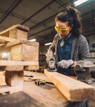 A middle-aged woman wearing a denim shirt, sweater, safety glasses and gloves cuts a 2x4 with a powersaw.  Cut wood is stacked carefully beside her.