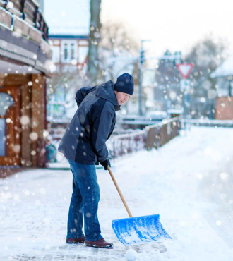 Man in gray and black coat with ski cap shovels the cobblestone walkway in front of a business.