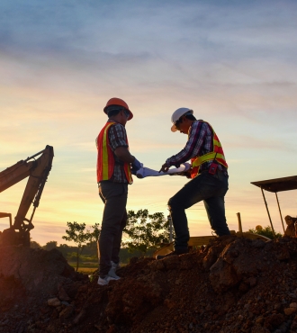 Two construction workers at a job site. They are wearing PPE and there is equipment and a sunset in the background.