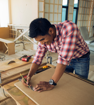 A contractor in a red plaid shirt marks a measurement on a sheet of particle board.