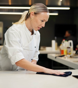 A blond female in a white chef coat wipes up the counter of a commercial kitchen. 
