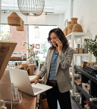Woman on a phone looking at a laptop behind the counter at a small retail store. 